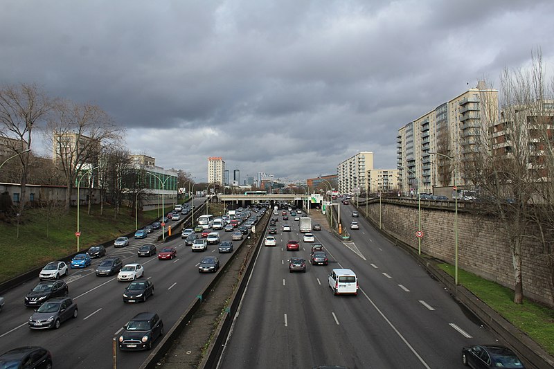 Highway with four lanes in each direction, busy but moving. A slip road exits on the right, with another entering on the left; on the right is a residential tower block