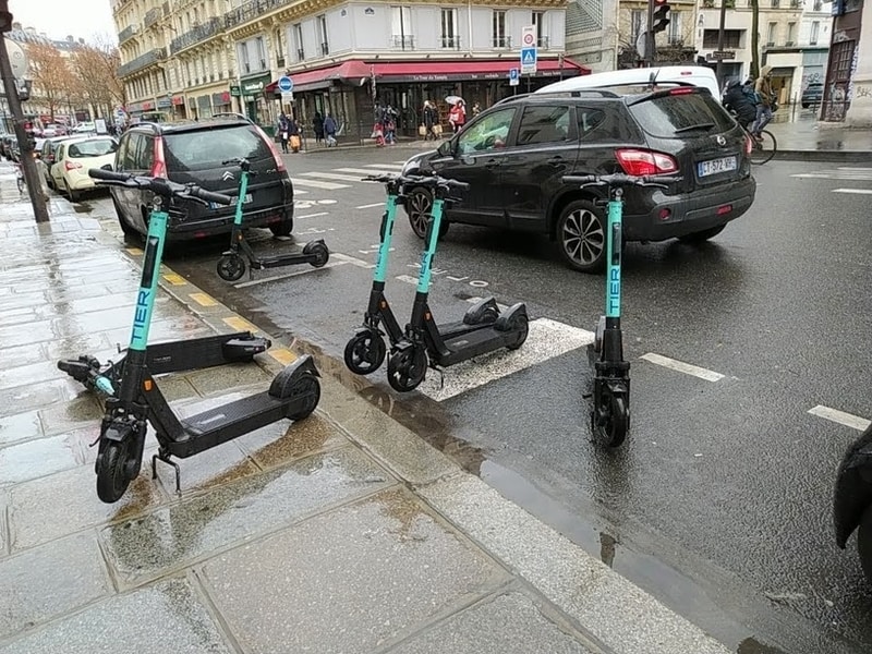 Electric scooters parked at a roadside. A car occupies the designated scooter parking spot, with scooters in the adjacent cycle and car parking spaces