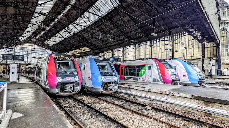 Modern single-deck trains in a terminus station. Two are pink, grey and white, with prominent SNCF logos; the other two are blue, grey and white, and feature both SNCF and Île-de-France Mobilités logos