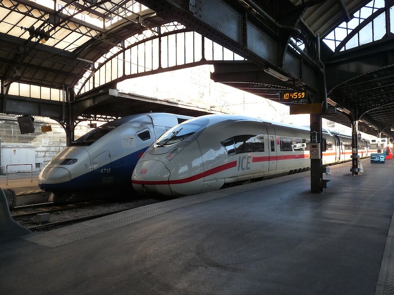 Two pointy-nosed high-speed trains. In the foreground, a white train with DB logo on the front and ICE on the side, along with a red line. Behind it, a grey and blue train