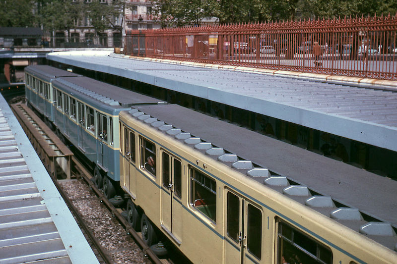 View of metro train from above in a station in a trench. Two blue cars and one cream car are visible