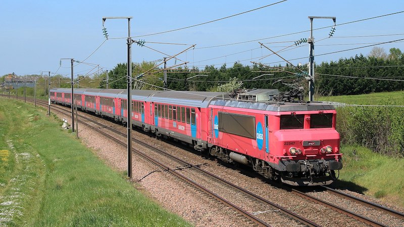 Distinctive BB 22347 electric locomotive with small SNCF logo on front, pulling Corail passenger cars, all in a pink livery with blue circles labelled Ouigo