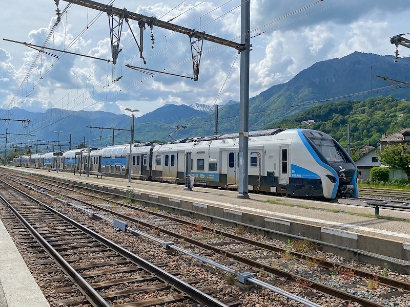 White, blue and grey train with single-deck end carriages and double-decker cars in between. Green mountains form the backdrop