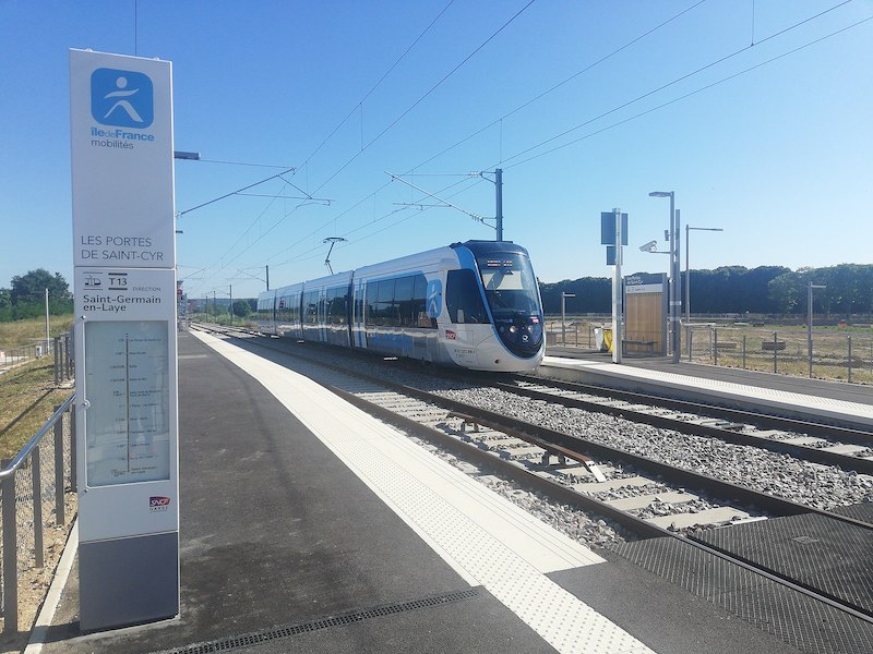 Train in white, blue and grey, sporting a small SNCF logo, at a tram platform surrounded by trees and fields