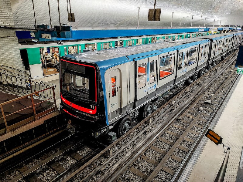 Two trains either side of an island platform. The first, in the foreground, is a modern walk-through train in blue, white and grey Île-de-France Mobilités livery. The second, in the background, is a much older train in green and white RATP livery