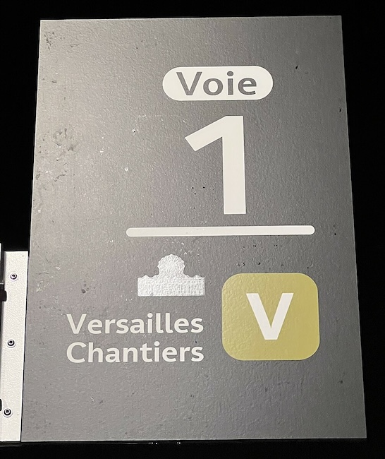 Sign reading Voie 1 : Versailles Chantiers, with the letter V in a rounded green-brown square