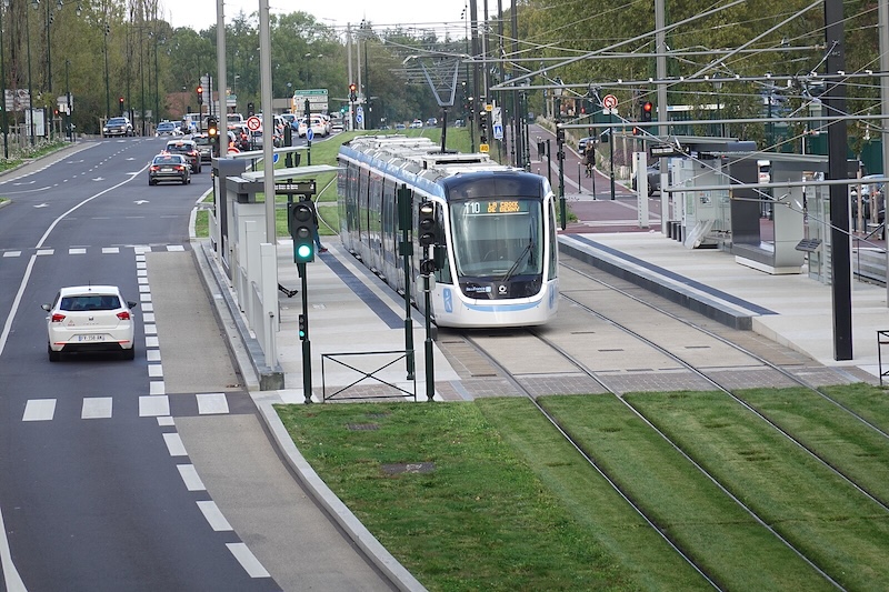 Modern tramway in white, blue and grey livery, stopped at a station. Outside the station, the tracks are planted with grass