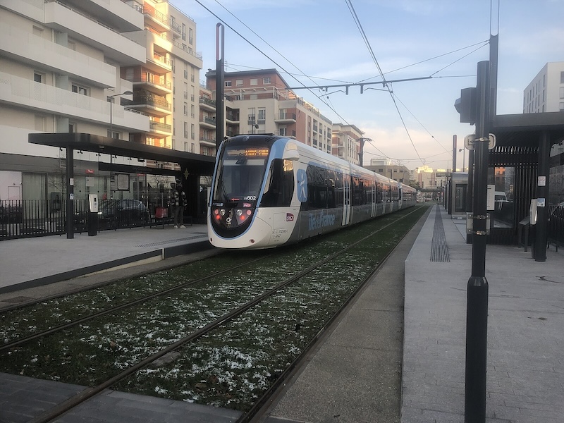 Tram stop planted with grass with some snow. An Alstom Citadis Dualis tram-train stands at the platform. Mid-rise buildings can be seen in the background