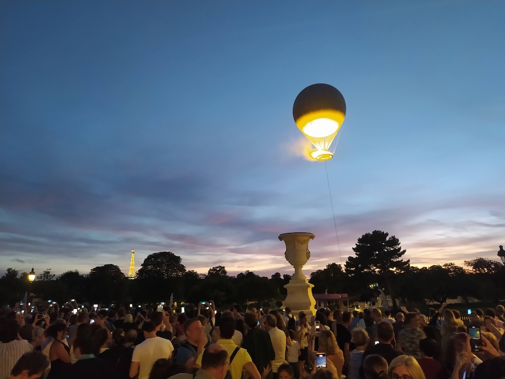 Tethered balloon with Olympic flame. A crowd is gathered in the foreground. In the distance, the Eiffel Tower is sparkling