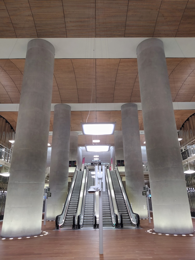 High wooden ceiling supported by concrete pillars. Ahead is a bank of 4 escalators