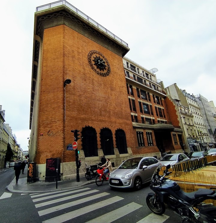 Red-brick wall with no adornments except a large iron clock near the top, and three iron lattices at street level. To the right, the building continues with windows.