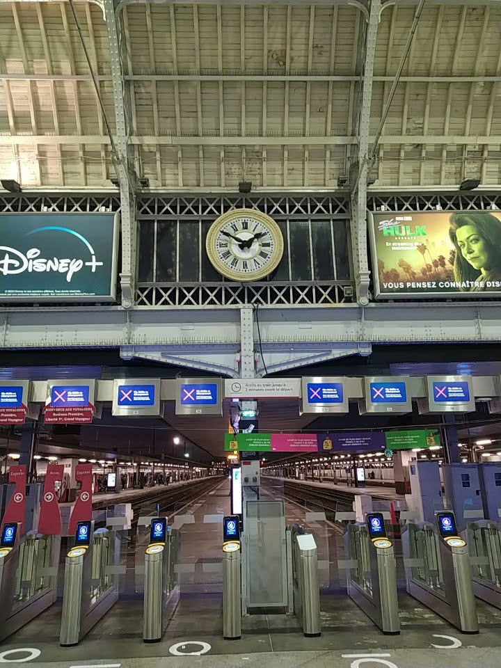 A row of ticket barriers. Above them, between two advertisements, is an old-fashioned analog clock, showing 1:50