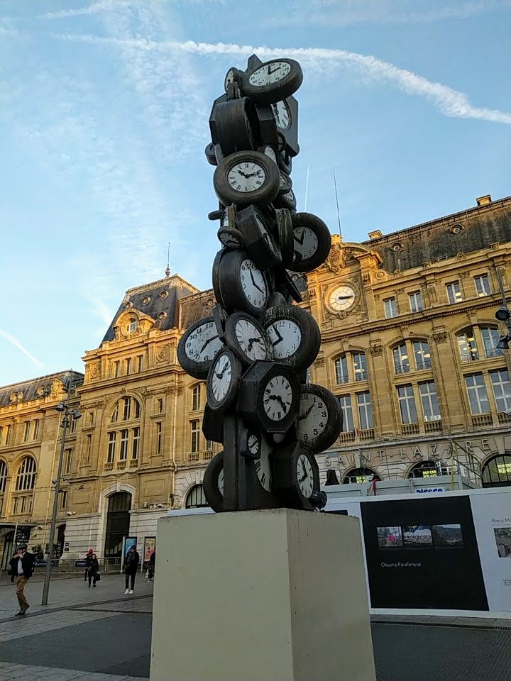 L'heure de tous sculpture. In the background, on the front wall of the Gare Saint-Lazare, a clock shows 3:15.