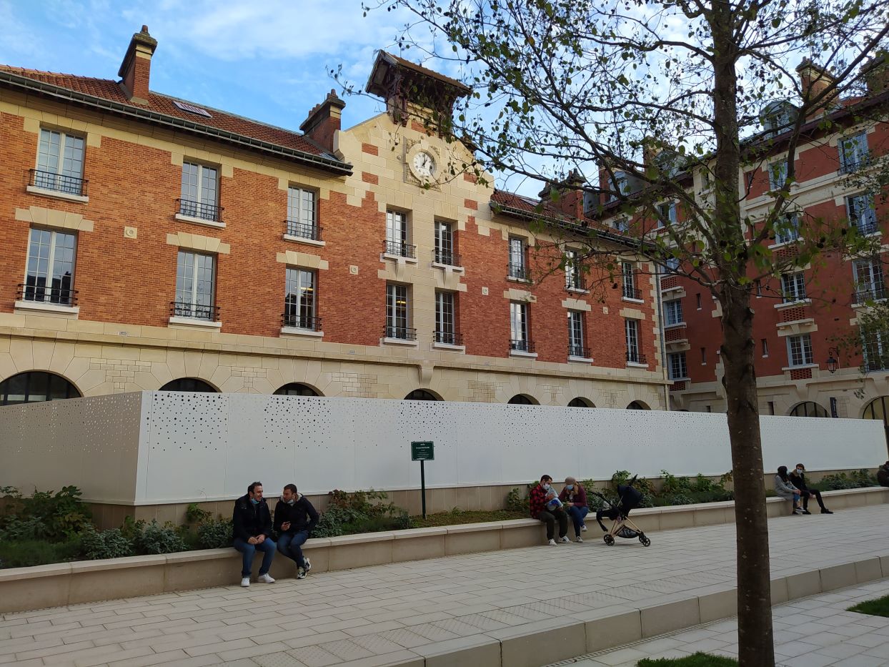 Clean red-brick buildings around a courtyard. The façade of one features a small tower with a clock. In front of this, small groups of people sit and chat, some of them masked