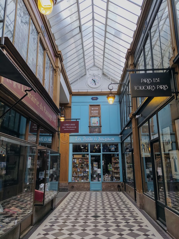 Analogue clock showing 10:27. Above is plasterwork reading 1846, and then a pitched glass roof; below is a brightly-lit, colourful passageway.