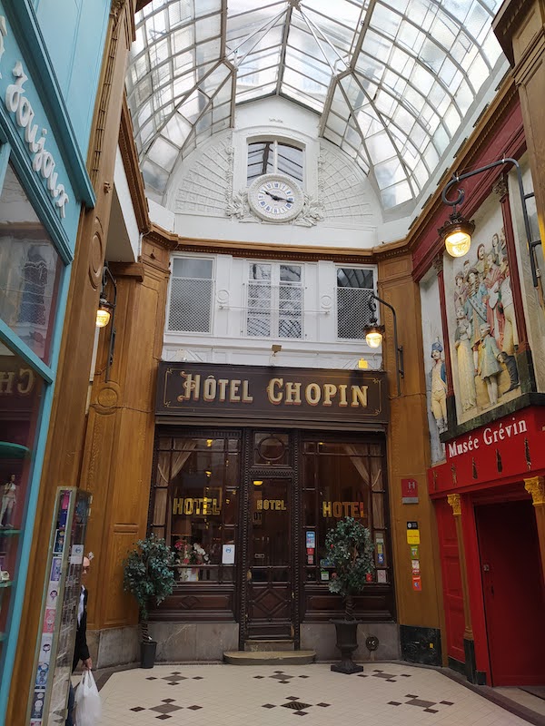 Analogue clock showing 10:17. Set below a window beneath an arched glass roof. The sign on the shopfront below reads Hôtel Chopin. On the right is an entrance to the Musée Grévin