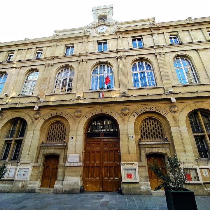 Stone building with arched windows. Above three arches at ground level are the words LIBERTÉ, ÉGALITÉ and FRATERNITÉ. Under the central arch, above large wooden double doors, are the words MAIRIE DU 2E ARRONDISSEMENT. Directly above this, at the top of the 3-storey building, is a clock showing 1:50