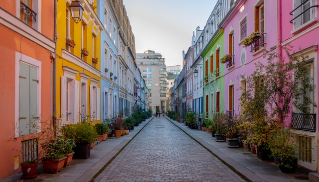 A paved street, empty of cars, lines with colourful 3-storey terraces fronted with plants. Two people stand together at the end of the road