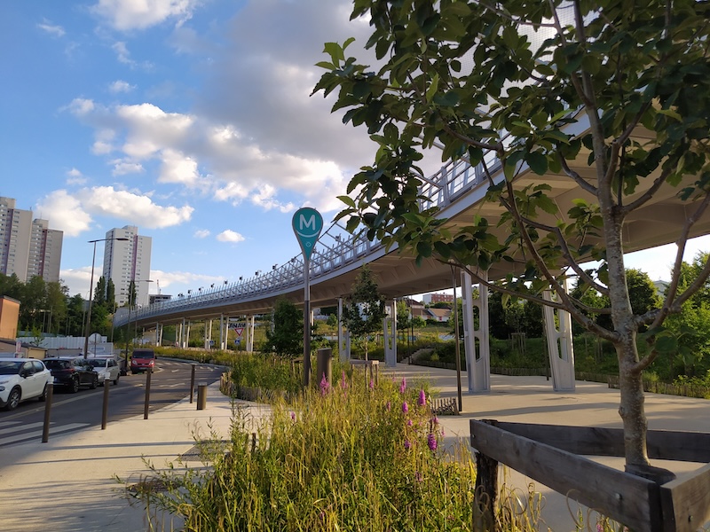 View from ground level of a viaduct extending into the distance, where several residential towers can be seen. A sign shaped like a map pin displays the M symbol of the metro and a small RATP logo