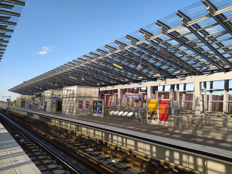 Metro platform, viewed from the opposite platform. Signs read Coteaux Beauclair. A glass canopy covers both platforms, with the tracks under the open blue sky.