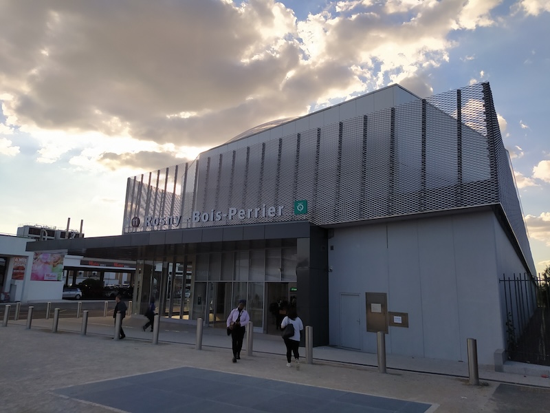 Box-shaped building, with a large glass entrance area on the left under a black canopy. At the upper level is a sort of wire mesh, in front of which is the name of the station in white lettering, between the coloured indicator of line 11 and the RATP logo.