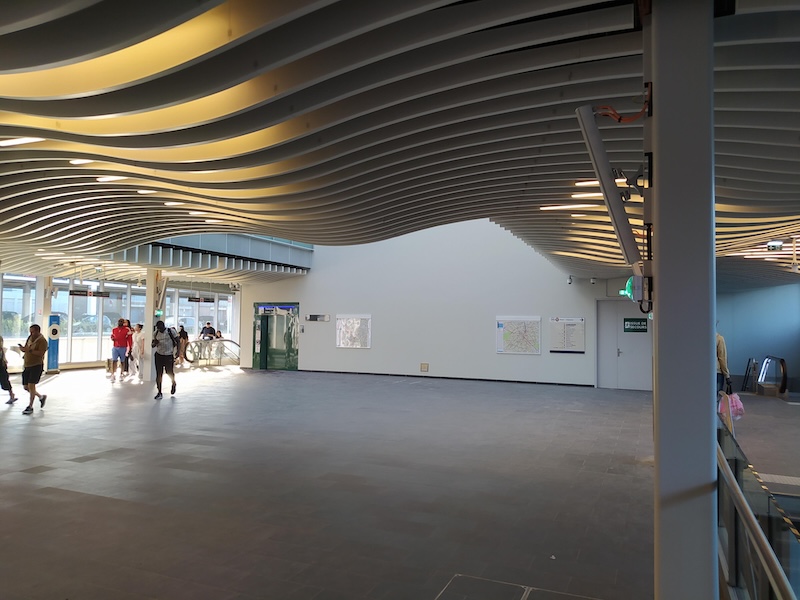 Open, square-shaped hall with white walls and wavy white ceiling decoration. Escalators lead down in the far corners. An automated toilet unit is visible next to the left-hand escalators. The left-hand wall is made of glass