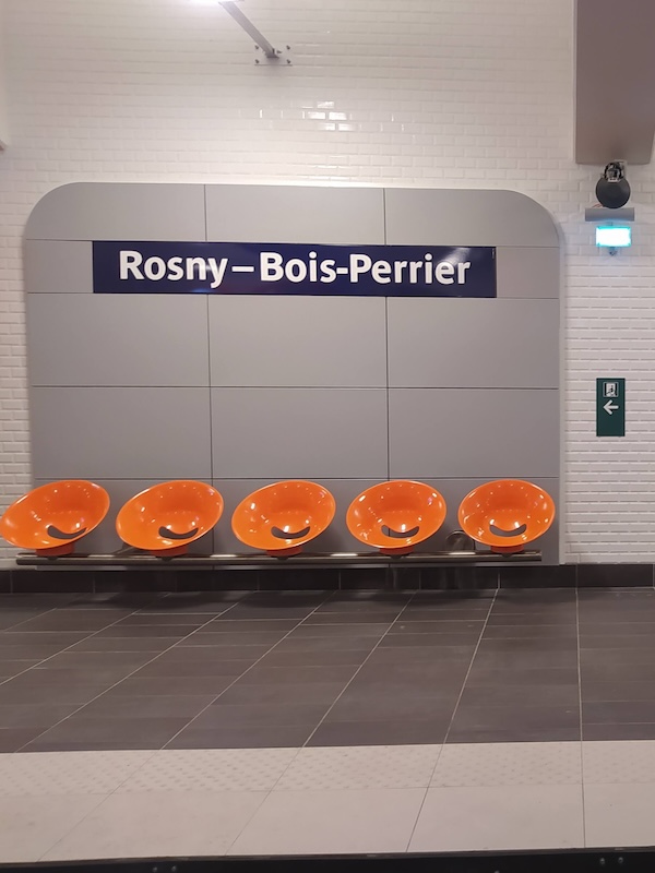 Metro platform, with grey floor tiles and bevelled white wall tiles. A grey plastic board is fixed to the wall, displaying the name of the station in white lettering on a blue background, and holding 5 round orange seats