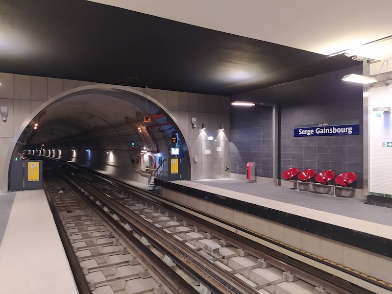 View of an underground metro platform. A sign reads Serge Gainsbourg over four red round chairs. An arched tunnel extends away from us