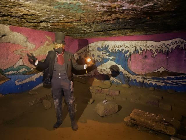 Man in top hat with lantern, in front of wave painting under a low earth ceiling