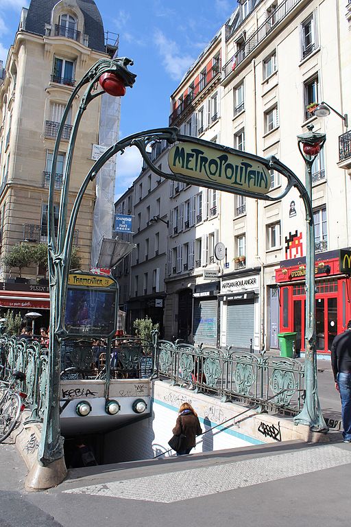 Art Nouveau metro entrance. In the foreground is an arch with the word METROPOLITAIN in curly lettering. Behind it, stairs lead downwards. Above them in the background is a sign with the station name PARMENTIER in the same lettering