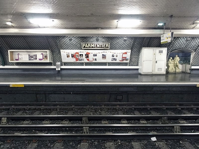 Metro station platform with white tiled wall, clad with a green trellis with a black and yellow sign reading PARMENTIER. Below the sign is a series of posters, and to the right, a statue