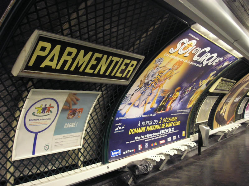 Black enamel sign with yellow lettering, reading PARMENTIER, against a green trellis. Next to the sign is an advertising poster, with white plastic tractor-style seats in front