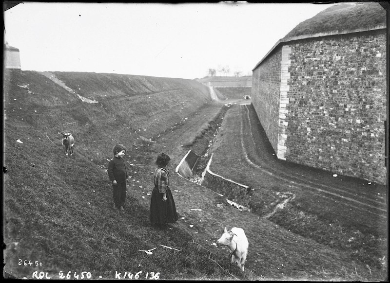 Black and white photograph of defensive wall, with children and goats in the adjacent grassy ditch