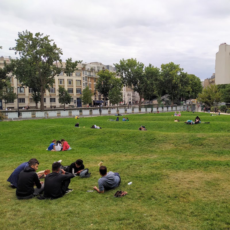 Groups of people sitting on a gently rolling lawn, with a canal in the background