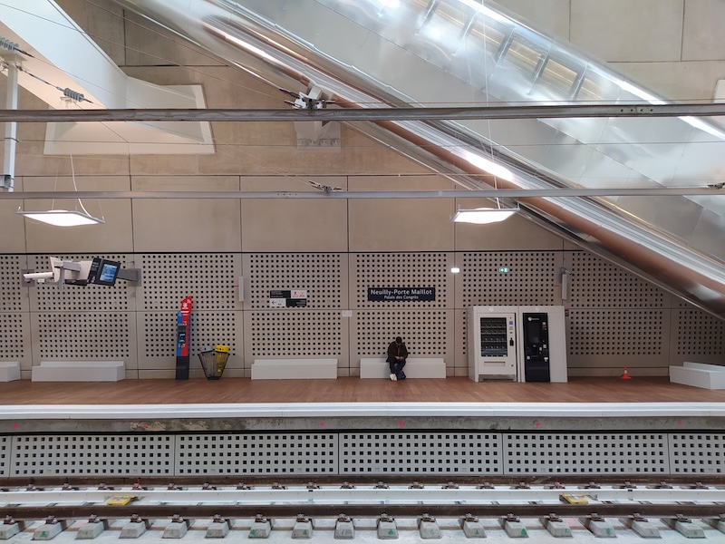 A passenger waits on a bench on a station platform. The sign above their head reads Neuilly - Porte Maillot (Palais des Congrés). Way above is an escalator