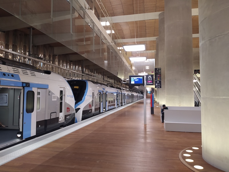 Double-decker train in white, blue and grey livery, standing at a platform at La Défense