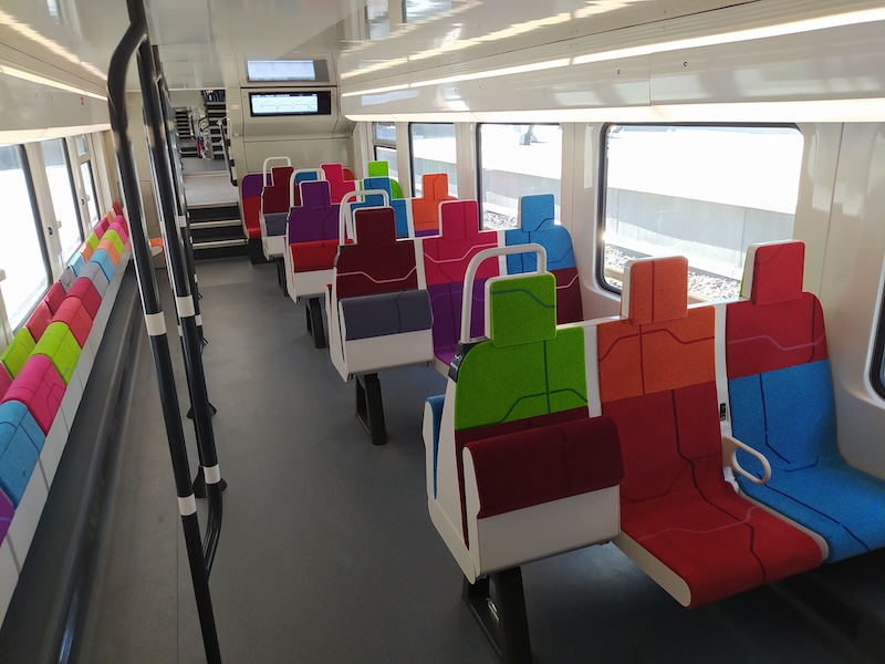 Train interior. On the left: sideways fold-down seats, and poles for standing passengers. On the right: transverse rows of three seats