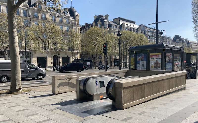 Low wall with escalator leading underground. In the background are cars and traditional Parisian buildings