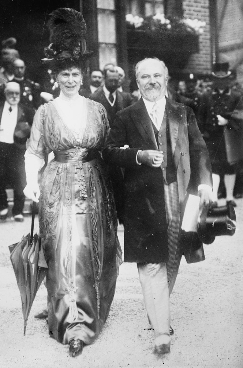 Black and white photograph of lady in high hat, arm in arm with bearded gentleman
