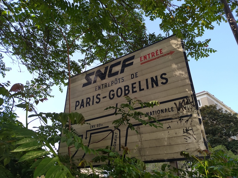 Old-looking sign, surrounded by overgrown vegetation, with an old SNCF logo, signalling the entrance to the ENTREPÔTS DE PARIS-GOBELINS