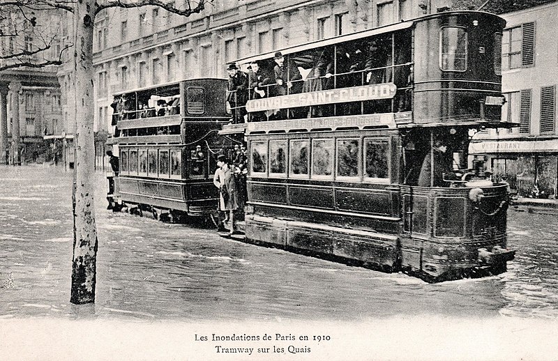 A two-car double-decker tram labelled LOUVRE – SAINT-CLOUD runs through shallow water. The postcard is labelled Les Inondations de Paris en 1910 / Tramway sur les Quais