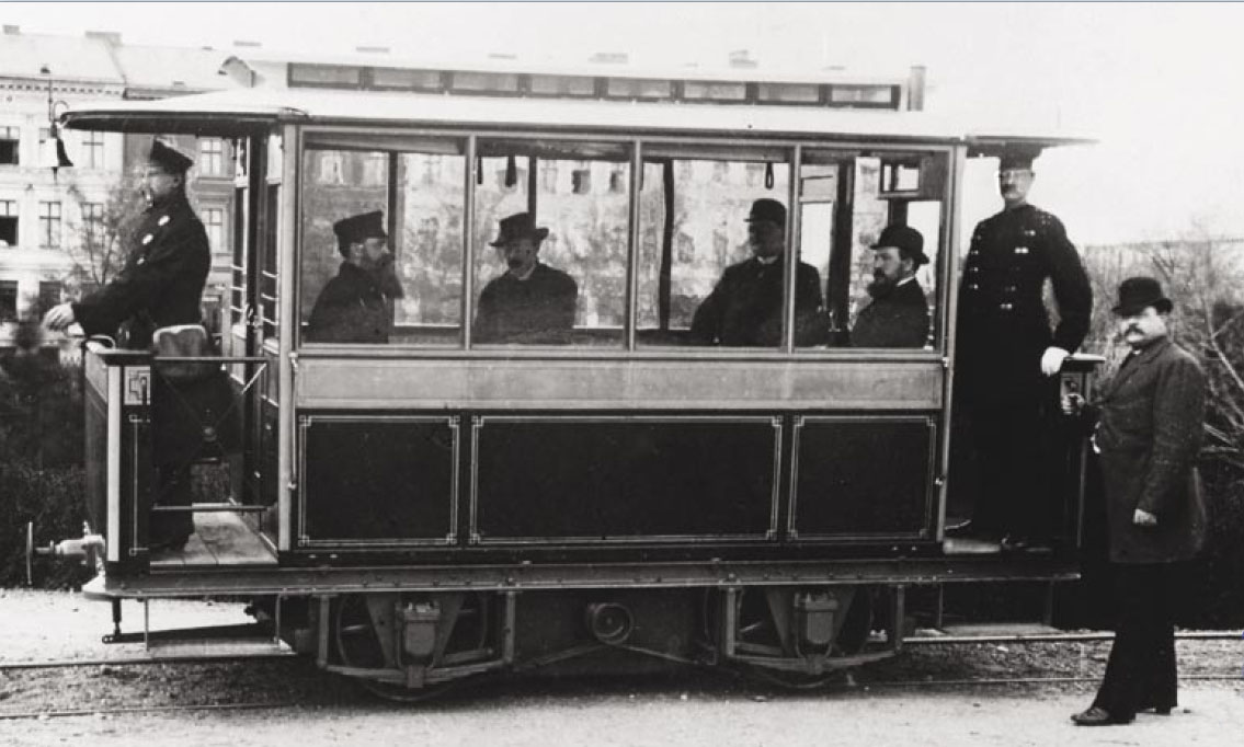 Black and white photo of a tram, carrying six smartly-dressed men, including one at each end in what looks to be a uniform. A seventh man stands to one side, looking at us