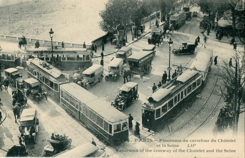 Street scene showing two two-car trams, one blocking the other. A dozen or so motor vehicles and a few horse-drawn carriages share the street, along with lots of pedestrians