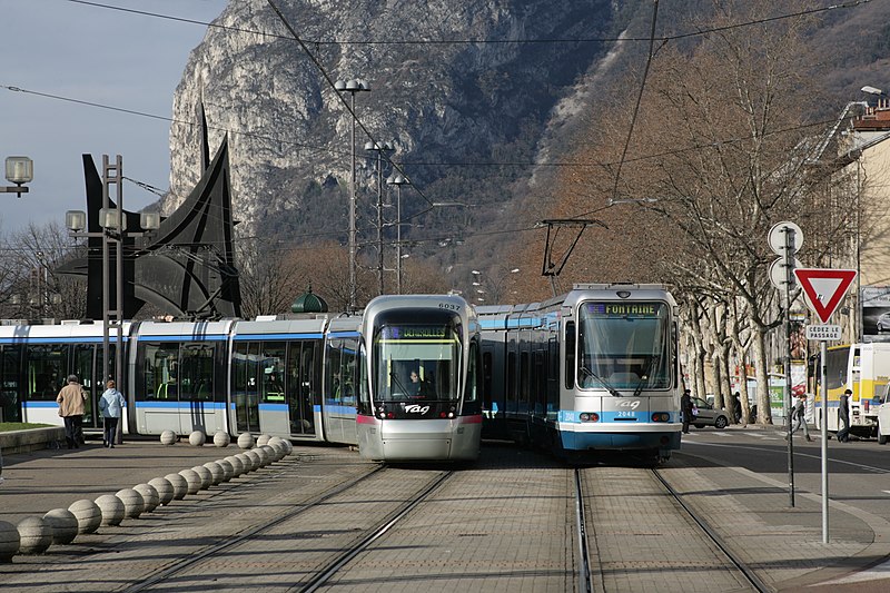Two trams pass in Grenoble: a modern Citadis and an older TFS. Both are liveried in silver and blue, with the TAG logo on the front. A mountain forms the backdrop