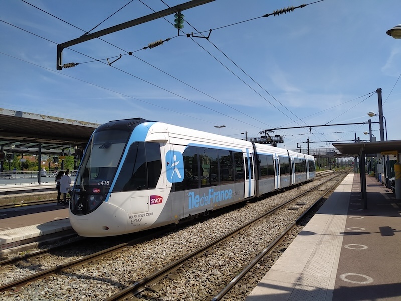 A tram-train in Île-de-France Mobilités livery (light blue, grey and white), standing in a station