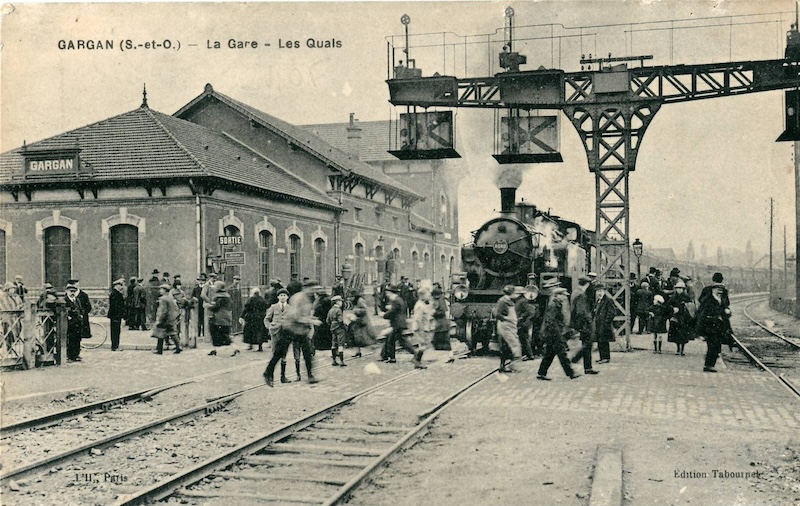 Sepia photo of a railway station. On the left is the building, with a sign reading GARGAN. On the right is a steam engine with steam bellowing from its chimney. Several dozen people move around on the platform and across the tracks. A gantry stands above the train