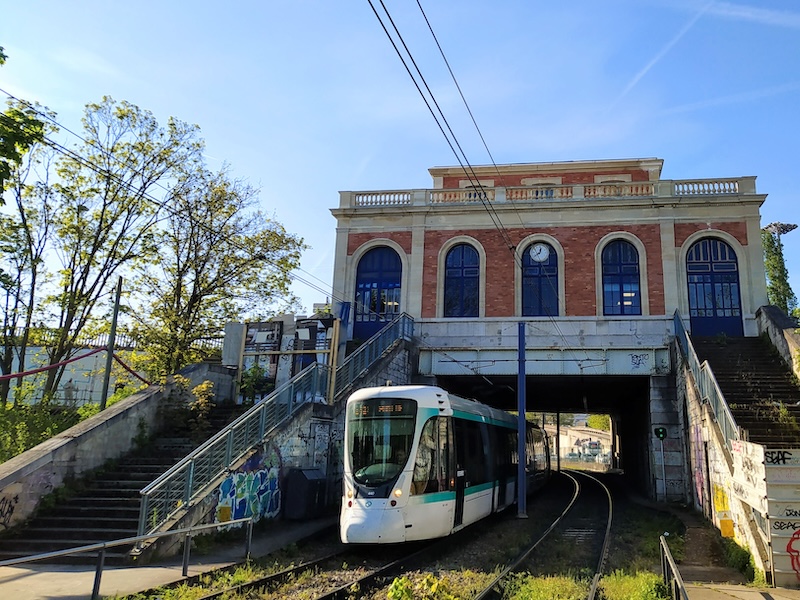 A tram passes along a railway under a red-brick building. Stairs leading to the building are blocked off, the barriers adorned with graffiti