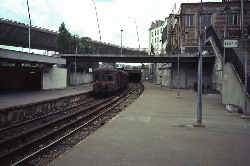 Colour photo of a grey scene taken from a railway platform. A train sits at the opposite platform. A concrete footbridge carries a single man over the tracks