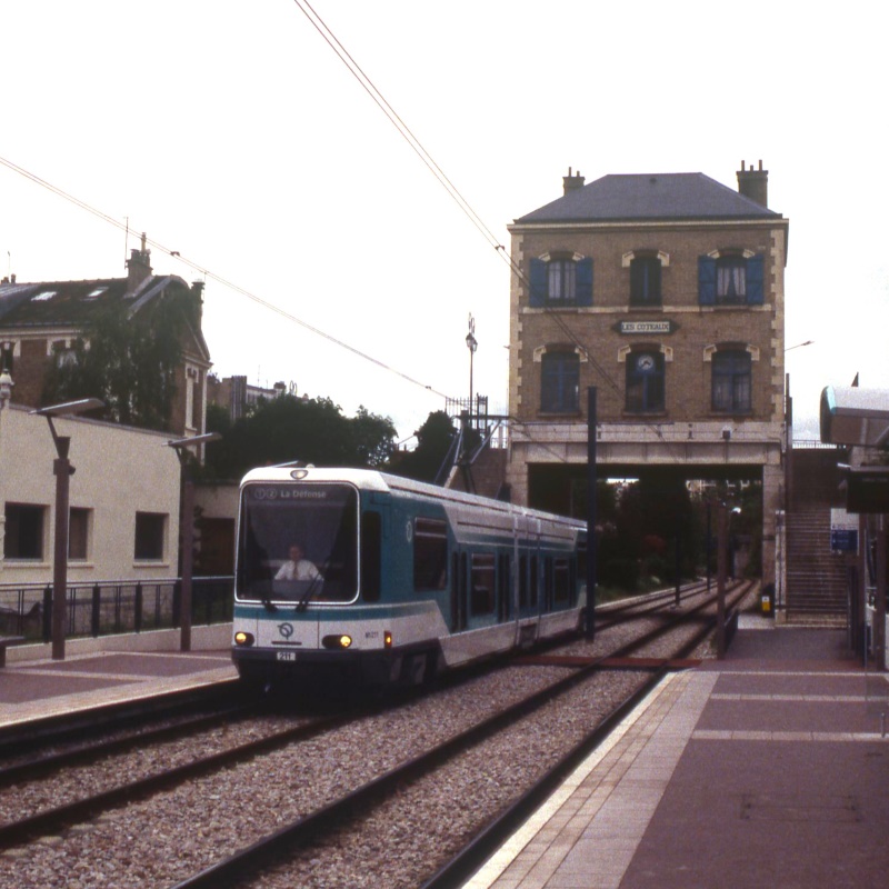Colour photo of a tram in a station. The tram is a TFS, an articulated vehicle with a high floor at either end but a low floor in the middle section. Behind the tram is a brick station building, standing over the tracks, with the name LES COTEAUX on a sign above the window.
